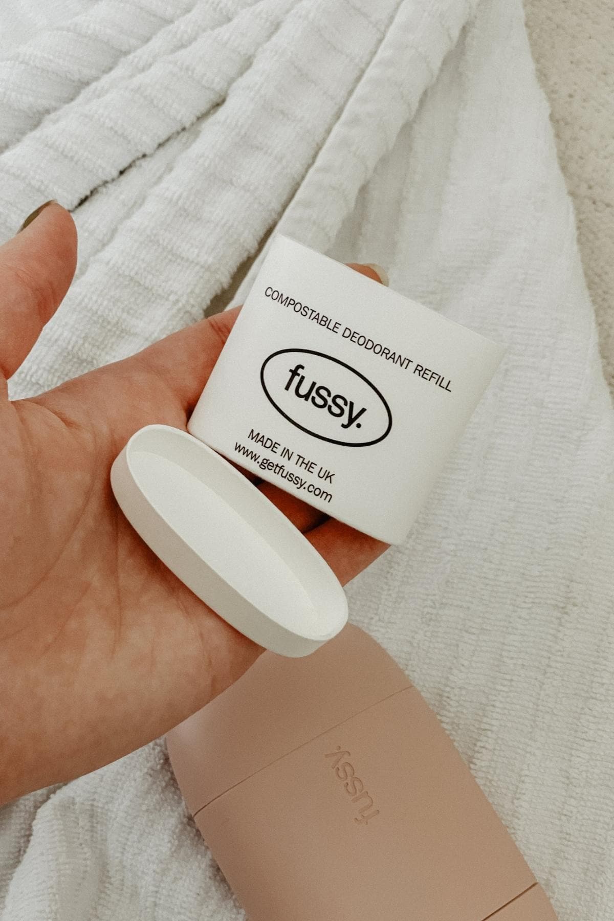A hand holding a compostable deodorant refill container of fussy deodorant with a removable lid, over a textured white surface. A pink deodorant case is visible underneath.