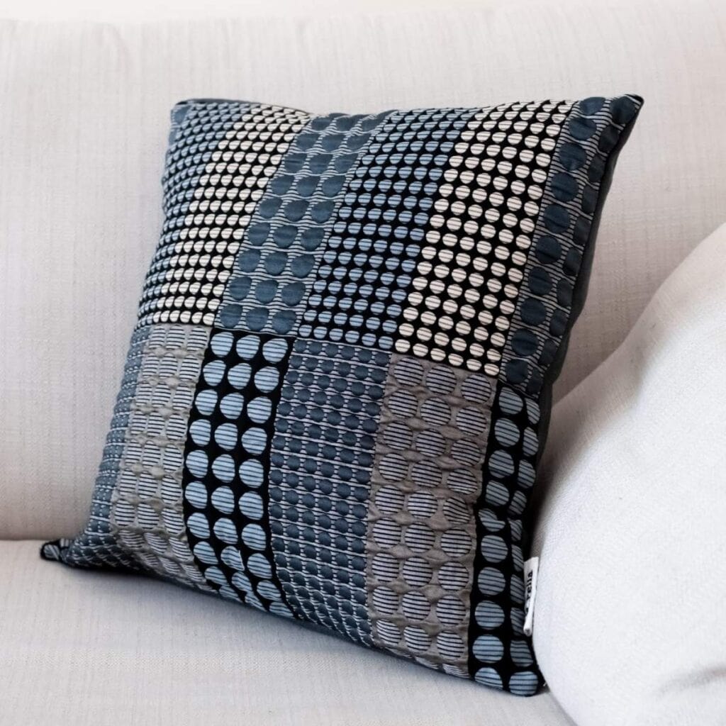 A decorative pillow with a patchwork design sits on a light-colored sofa, perfect as a handmade wedding gift. It features various shades of blue and gray with circular patterns, creating a textured look thats both stylish and budget-friendly.