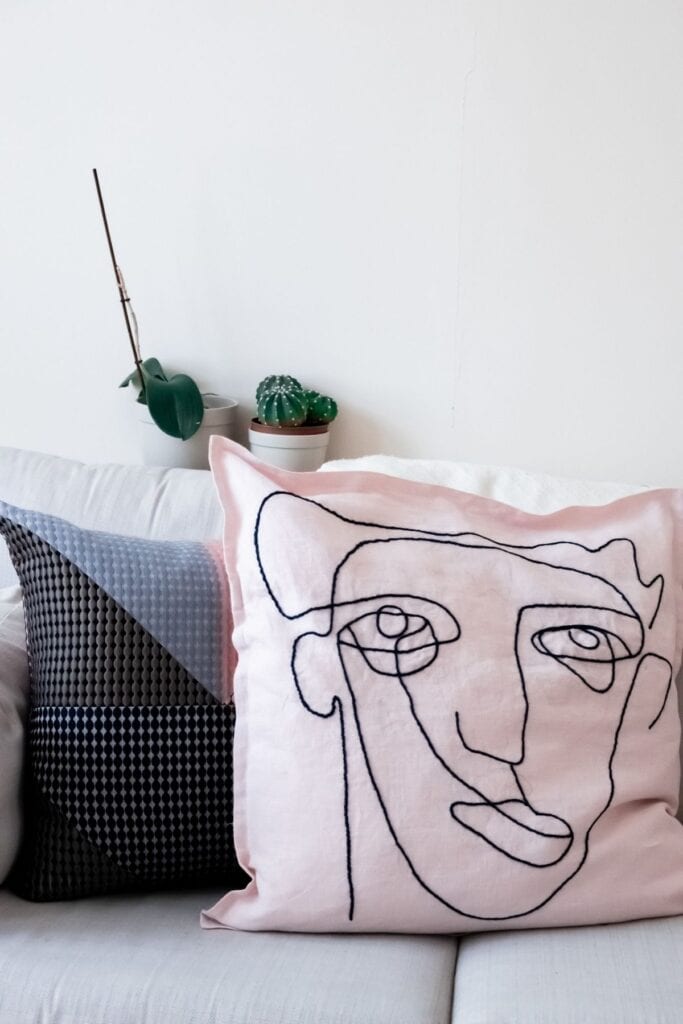 Two decorative pillows on a white sofa: one pink with a minimalist abstract face line drawing, and another with a geometric pattern. Perfect as handmade wedding gifts, they add charm to any space. In the background, a small potted plant and cactus are placed on a shelf.