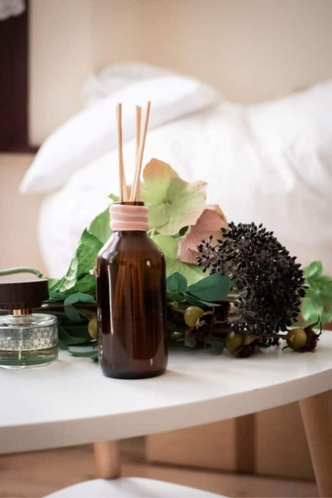 A brown glass bottle with reed diffusers graces a white table, surrounded by green leaves, pink and white flowers, and a black dried flower cluster. Perfect for handmade wedding gifts on a budget, it sits elegantly beside another small glass jar. A bed with white linens rests in the background.