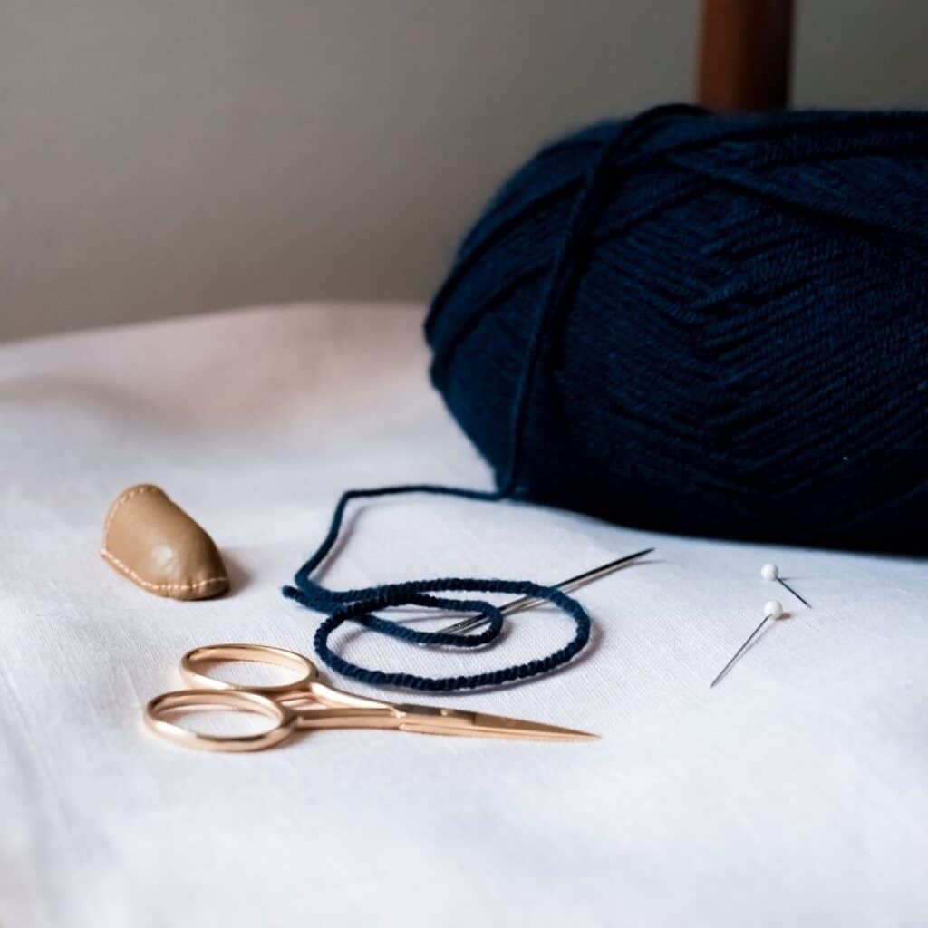 Close-up of a knitting and sewing setup on a white surface, perfect for crafting handmade wedding gifts. It features a ball of dark blue yarn, a needle, two pins, gold-colored scissors, and a leather thimble.