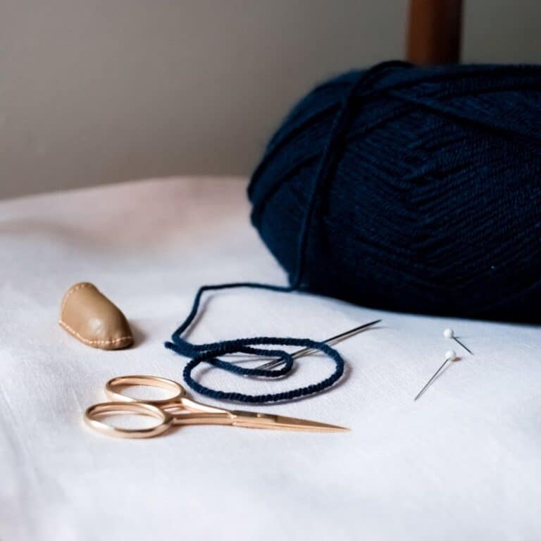 Close-up of a knitting and sewing setup on a white surface, perfect for crafting handmade wedding gifts. It features a ball of dark blue yarn, a needle, two pins, gold-colored scissors, and a leather thimble.