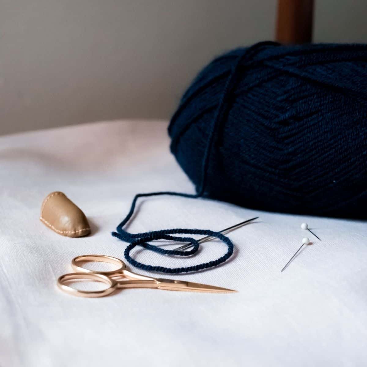 Close-up of a knitting and sewing setup on a white surface, perfect for crafting handmade wedding gifts. It features a ball of dark blue yarn, a needle, two pins, gold-colored scissors, and a leather thimble.