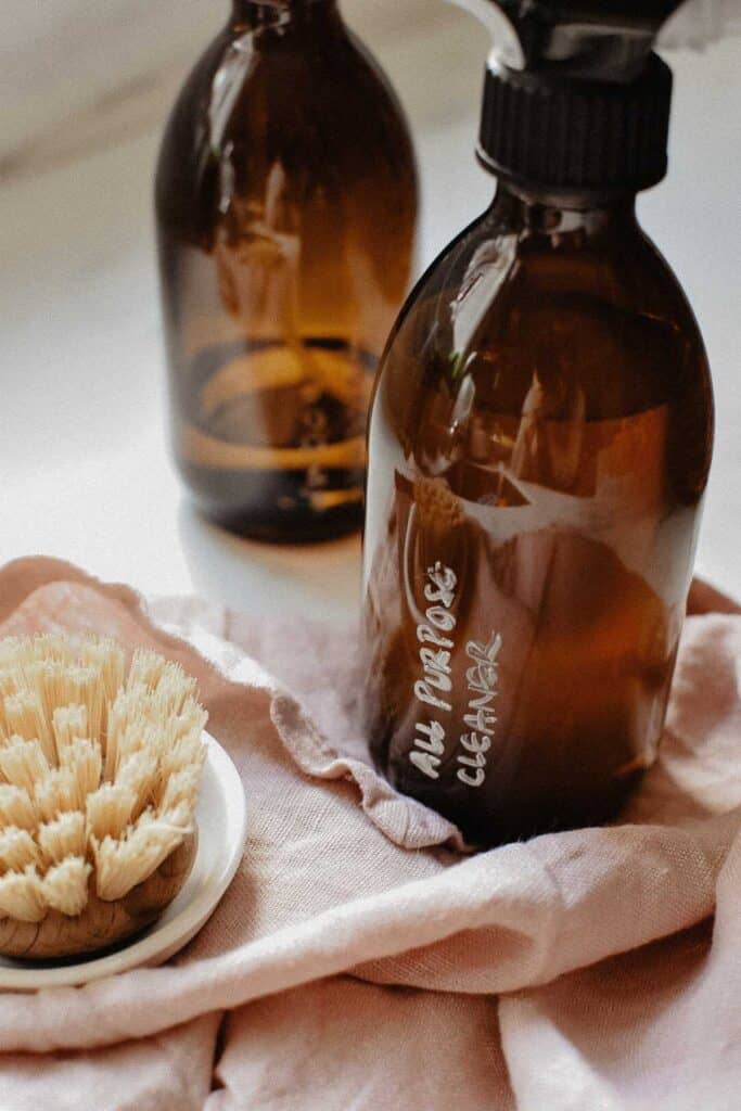 Two amber glass bottles labeled All Purpose Cleaner rest on a soft pink cloth, accompanied by a natural bristle brush. This serene scene of essential homemade cleaning products hints at an eco-friendly approach.