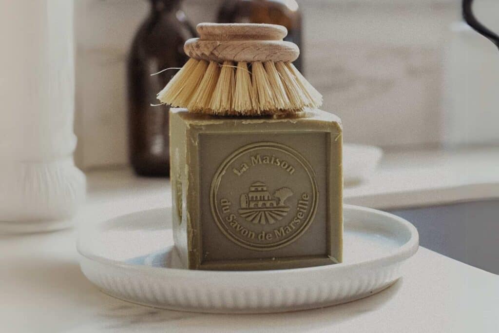 A square block of green soap, sits embossed with La Maison du Savon de Marseille on a white dish. 