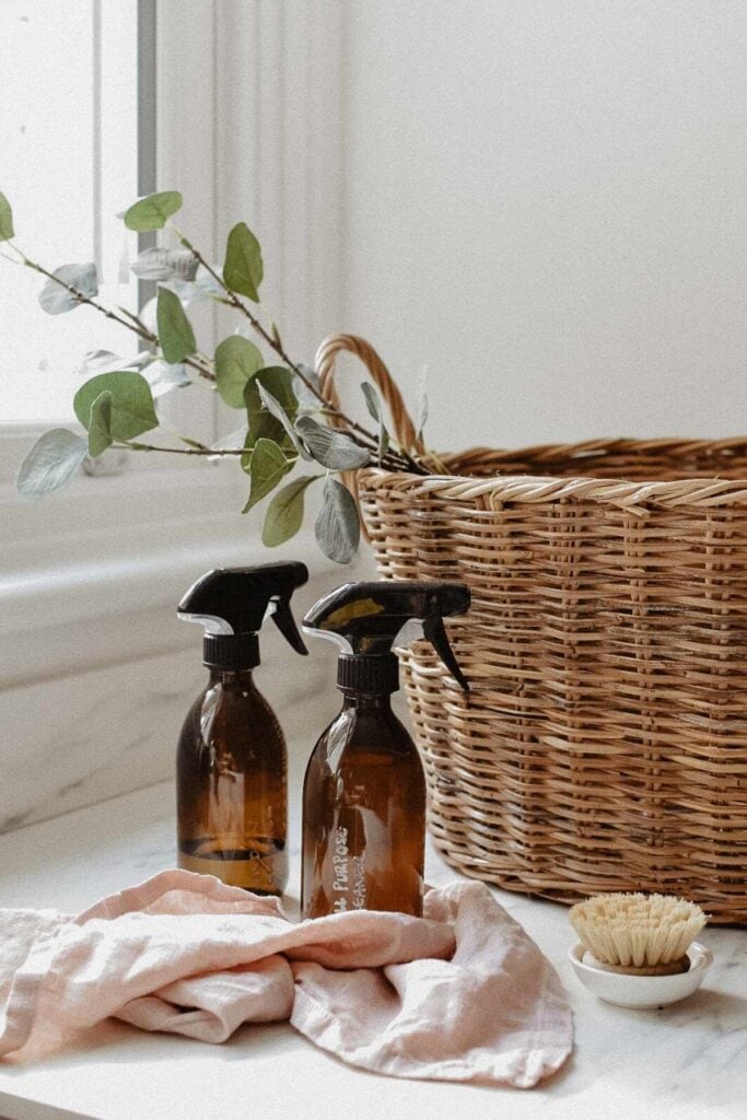 Two brown spray bottles with homemade cleaning products sit on a counter beside a beige cloth and a small brush. A wicker basket with greenery graces the background near a window. 