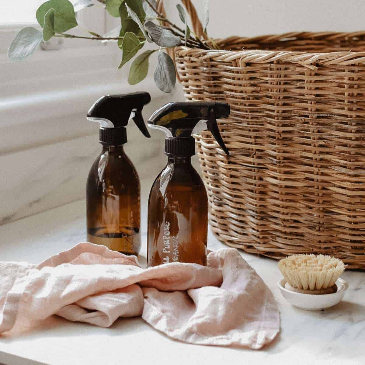 Two amber spray bottles sit on a countertop near a wicker basket and a dish brush, showcasing homemade cleaning products. A soft pink cloth is draped nearby, with green leaves partially visible in the background.