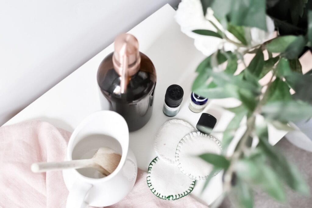 A white table with a pink cloth holds a white jug with a brush, a dark bottle, two small jars, and reusable facial pads, alongside ingredients for natural homemade face-wash. A large green leafy plant partially covers the view.