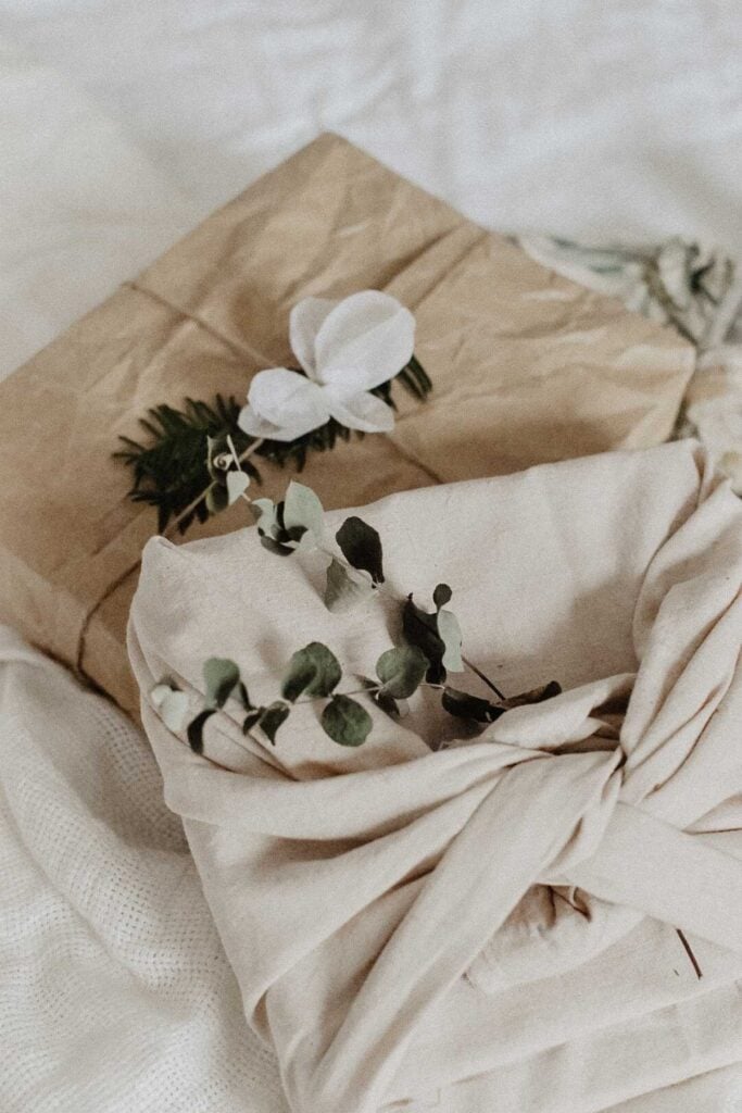 Two handmade gifts wrapped in fabric lie on a soft surface. The foreground package is elegantly wrapped in light beige with a knot, adorned with eucalyptus leaves, while the background package features brown paper topped with a white flower and greenery, perfect DIY gift ideas.