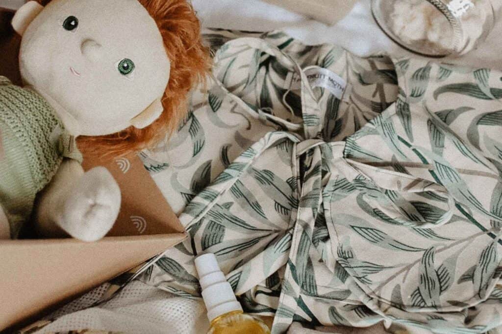 A soft fabric doll with red hair and a green outfit lies next to a leafy patterned blouse, perfect as handmade gifts. Nearby, theres a small spray bottle and a jar of cream, all on a light surface.