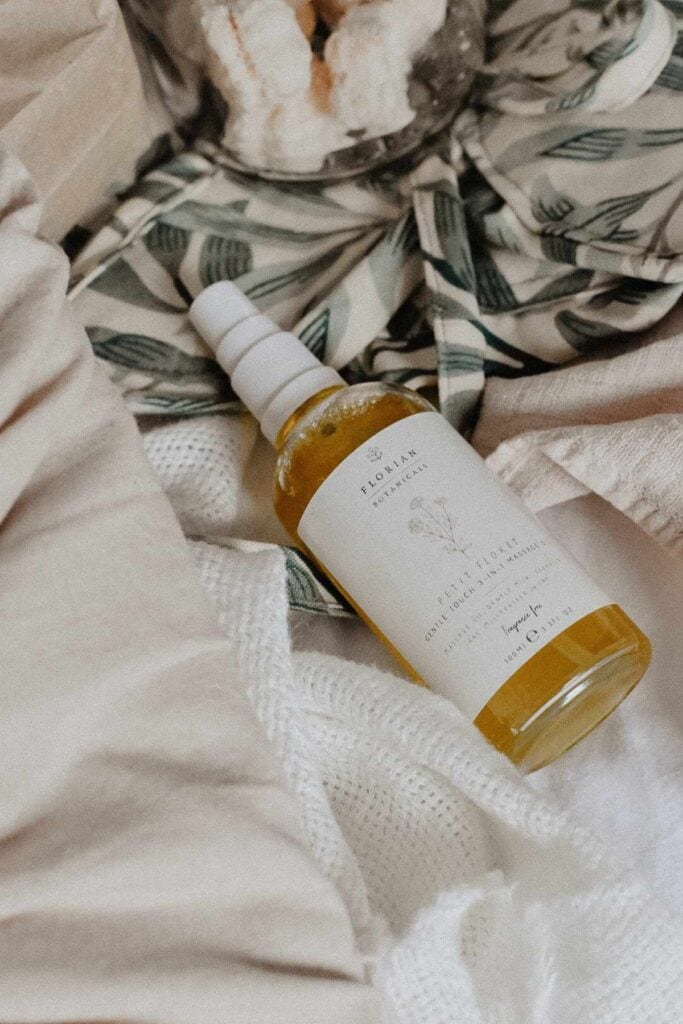 A bottle of face oil with a white label and a dropper rests on a textured fabric surface, ideal for those seeking DIY gift ideas. Nearby, a green leafy patterned cloth complements the scene, while in the background, a glass jar with homemade bath bombs hints at charming handmade gifts.