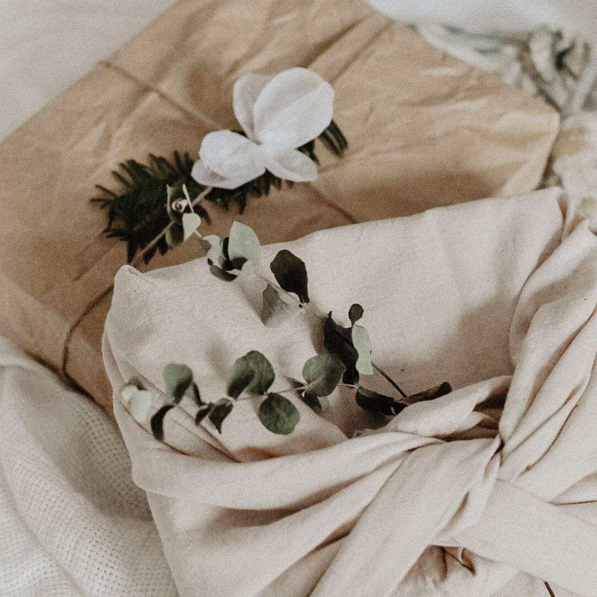 Two wrapped gifts are shown; one in brown paper adorned with a white flower and greenery, the other in a beige cloth tied with eucalyptus sprigs. Perfect for DIY gift ideas, theyre placed on a soft, white textured surface.
