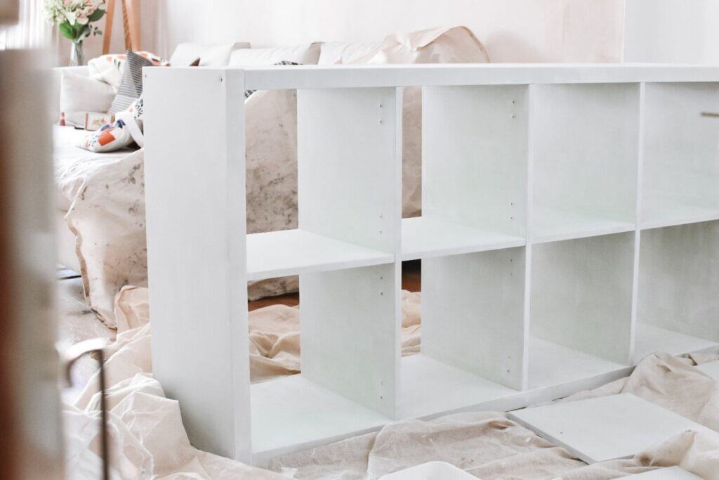 A primed IKEA Kallax is surrounded by protective sheets in the living room. Tools and cushions rest on the sofa in the background.