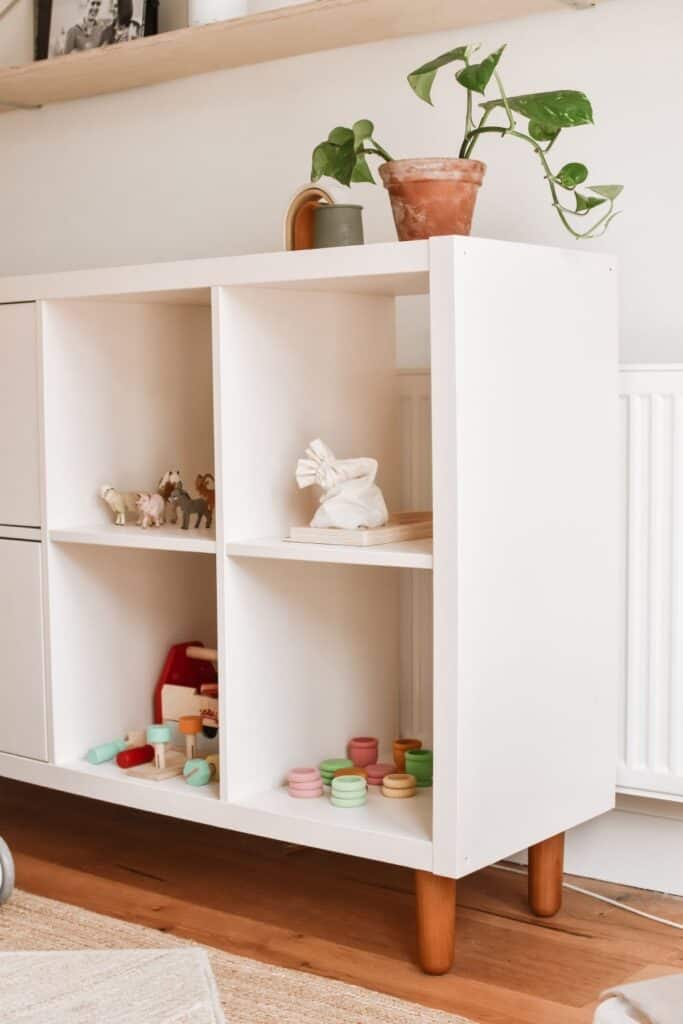 A pale pink shelving unit with four cubbies on wooden legs showcases a clever IKEA Kallax hack. It holds childrens toys, including animal figures and colorful cups. A potted plant and small jug sit on top, adding charm. 
