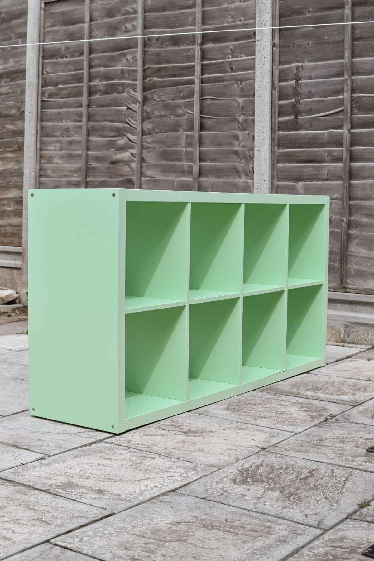 A DIY IKEA Kallax Hack begins with a mint green wooden cubby shelf featuring eight compartments, standing on a tiled outdoor patio. 