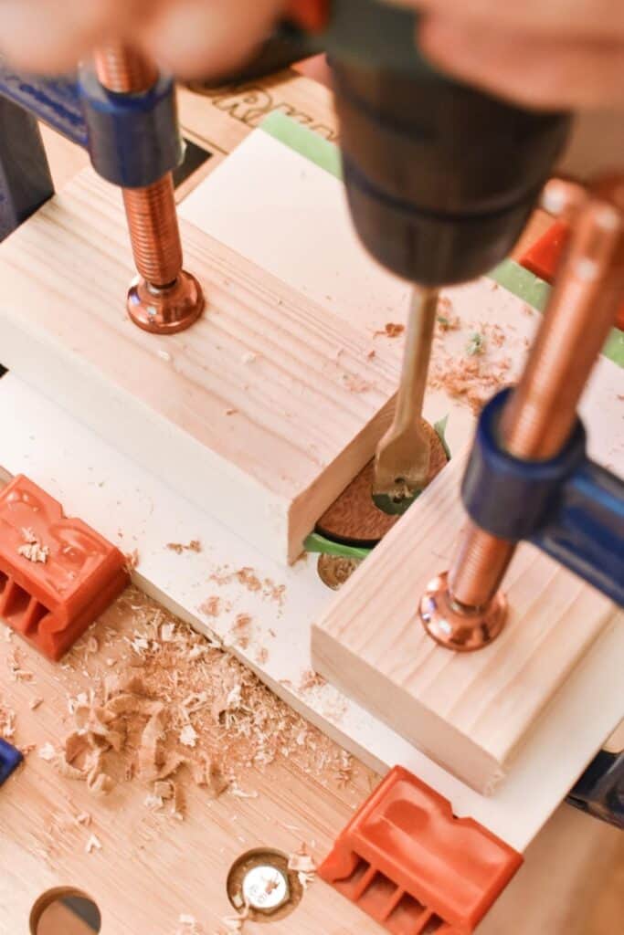 A close-up of a woodworking setup shows a drill making precise holes in a plywood shape, ready for adding to the IKEA Kallax hack. Two clamps keep the plank sturdy as wood shavings scatter around, while a green ruler rests in the background.
