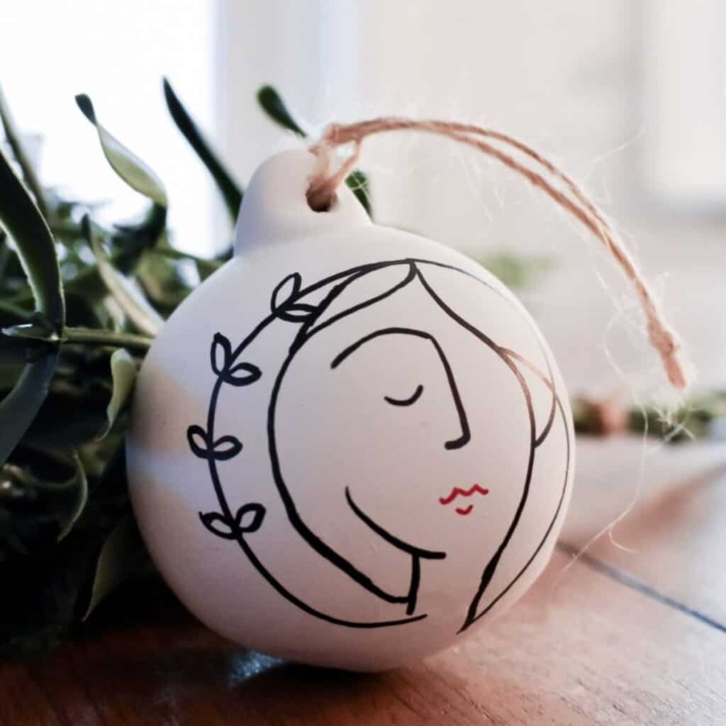 A ceramic ornament featuring a minimalist drawing of a womans face and vine rests on a wooden surface. With a brown string for hanging, this piece is perfect for those who love easy baubles. Green leaves are partially visible in the background.