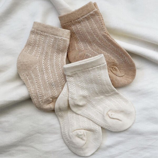 Four knitted socks in beige and cream, perfect as eco-friendly gifts for children or babies, are laid out on a white fabric. They feature a subtle chevron pattern, with two beige socks at the top and two cream ones at the bottom.