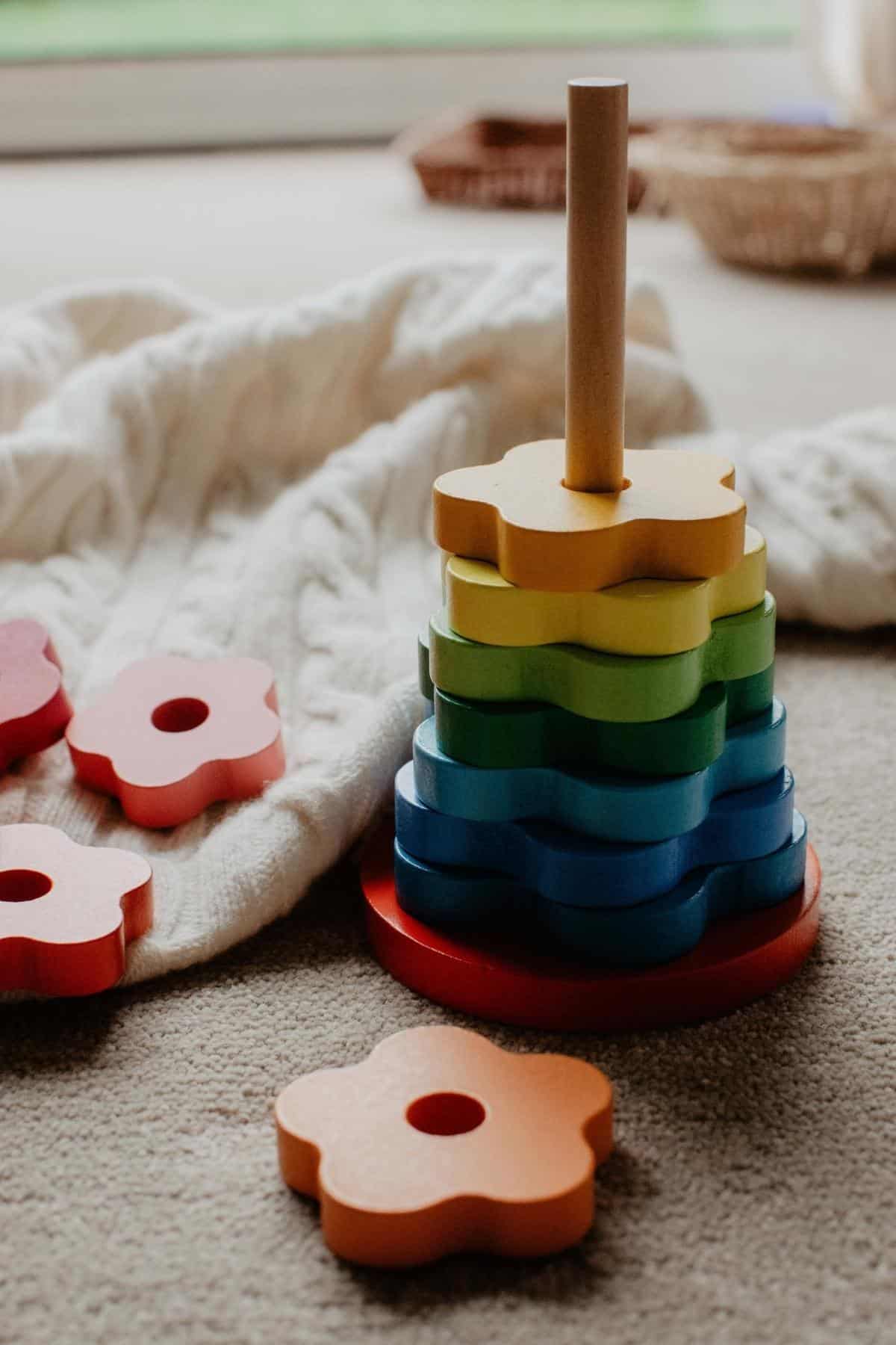 Colorful wooden stacking toy from Jaques of London, with flower-shaped pieces in vibrant hues like red, orange, yellow, green, and blue. Some pieces rest on a white knitted blanket nearby, all on a carpeted floor.
