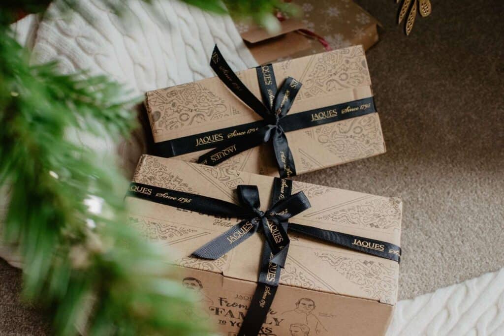 Two gift boxes wrapped in tan paper featuring Jaques of Londons illustrations are tied with black ribbon, resting on a beige carpet near a green Christmas tree branch. A white knitted fabric peeks through the scene, evoking memories akin to treasured wooden toys under the tree.