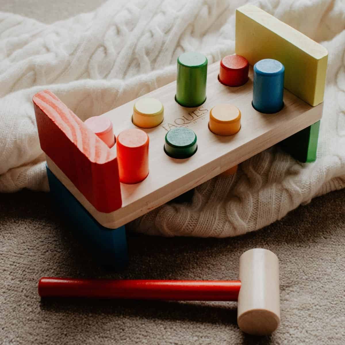 A charming wooden toy bench from Jaques of London sits on a soft, white knitted blanket. Nearby, a mallet with a red handle rests ready for play. The multicolored pegs—red, green, blue, and yellow—add vibrant fun thats classic in any wooden toys collection.