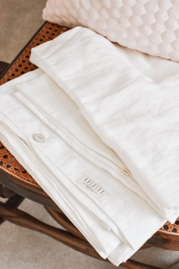 Folded white linen cot sheets with buttons are neatly placed on a wooden chair with a woven cane seat. A pink textured cushion is visible in the background, adding a soft touch reminiscent of delicate baby clothing.