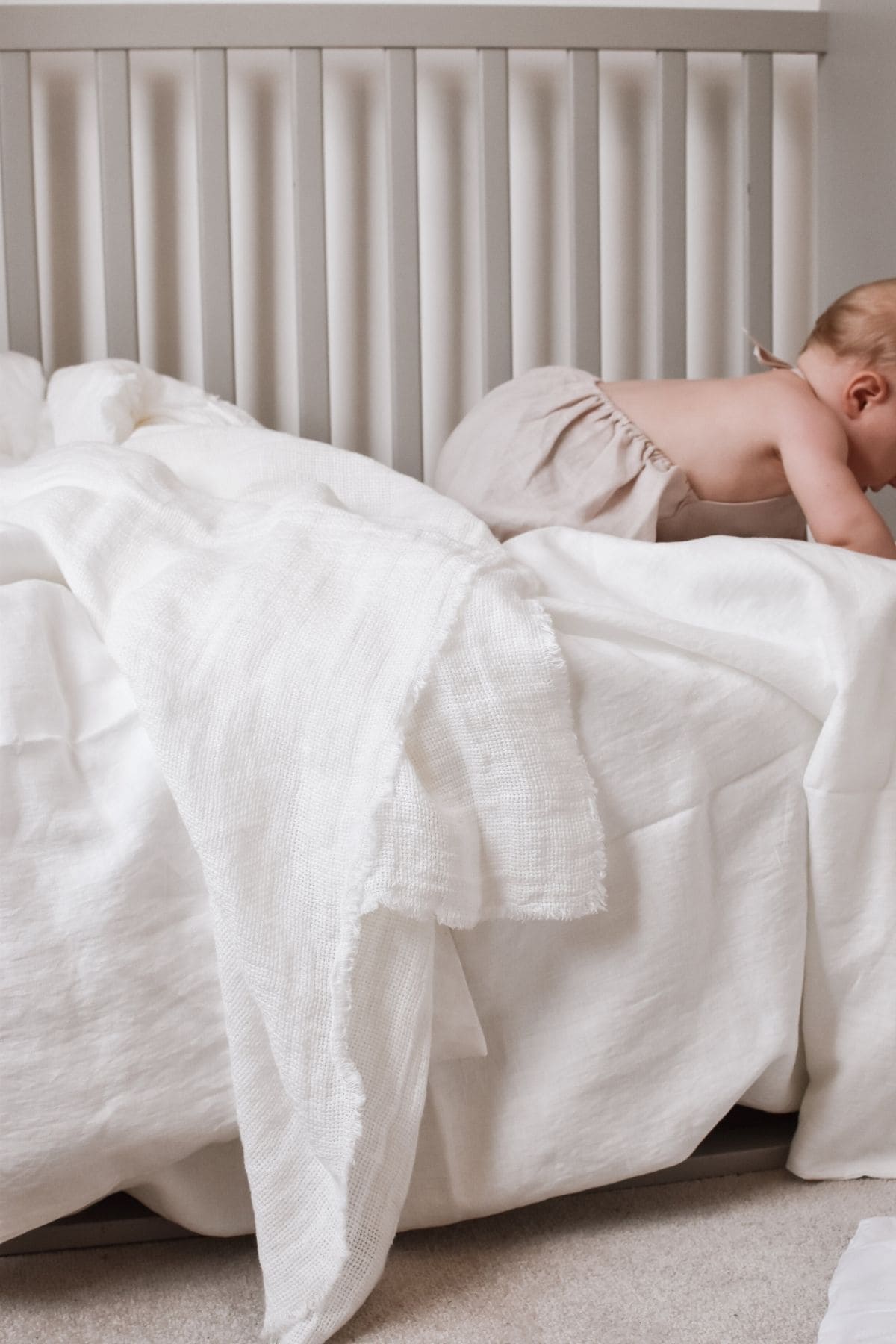 A baby in a beige outfit from Kalusha crawls on a white bed adorned with textured linen cot sheets. The cribs slatted headboard is visible in the background, adding to the serene nursery vibe.