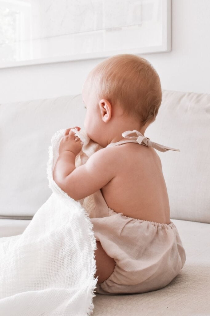 A baby in a light beige romper sits on a cream-colored sofa, cuddling a soft white blanket. The babys head turns slightly away, showing the back and profile.