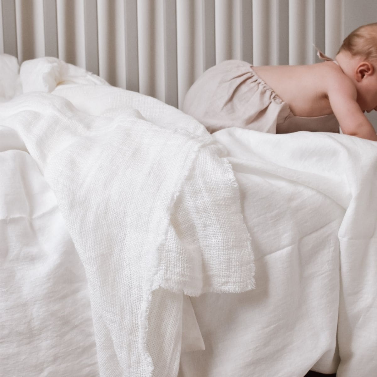 A baby dressed in Kalushas light-colored outfit is crawling on a bed layered with soft, textured linen cot sheets. Vertical blinds in the background contribute to the scenes gentle, neutral ambiance.