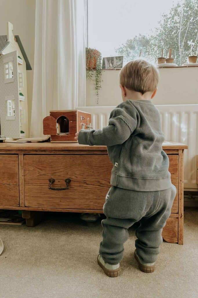 A toddler in a cozy green outfit is standing and playing with a wooden toy house on a wooden chest. A dollhouse and potted plants are visible nearby, with light streaming in from a window.