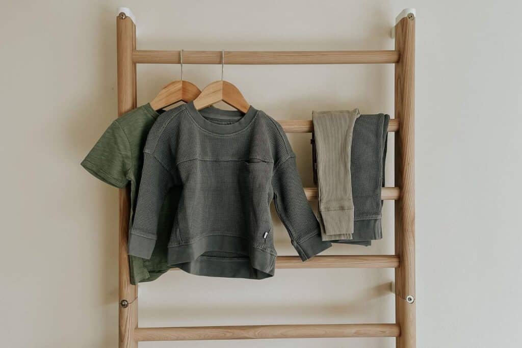 Two childrens long-sleeve Kidly shirts in green tones hang on wooden hangers from a wooden rack. Beside them, a pair of folded pants is draped over a rung. The background is a plain, light-colored wall.