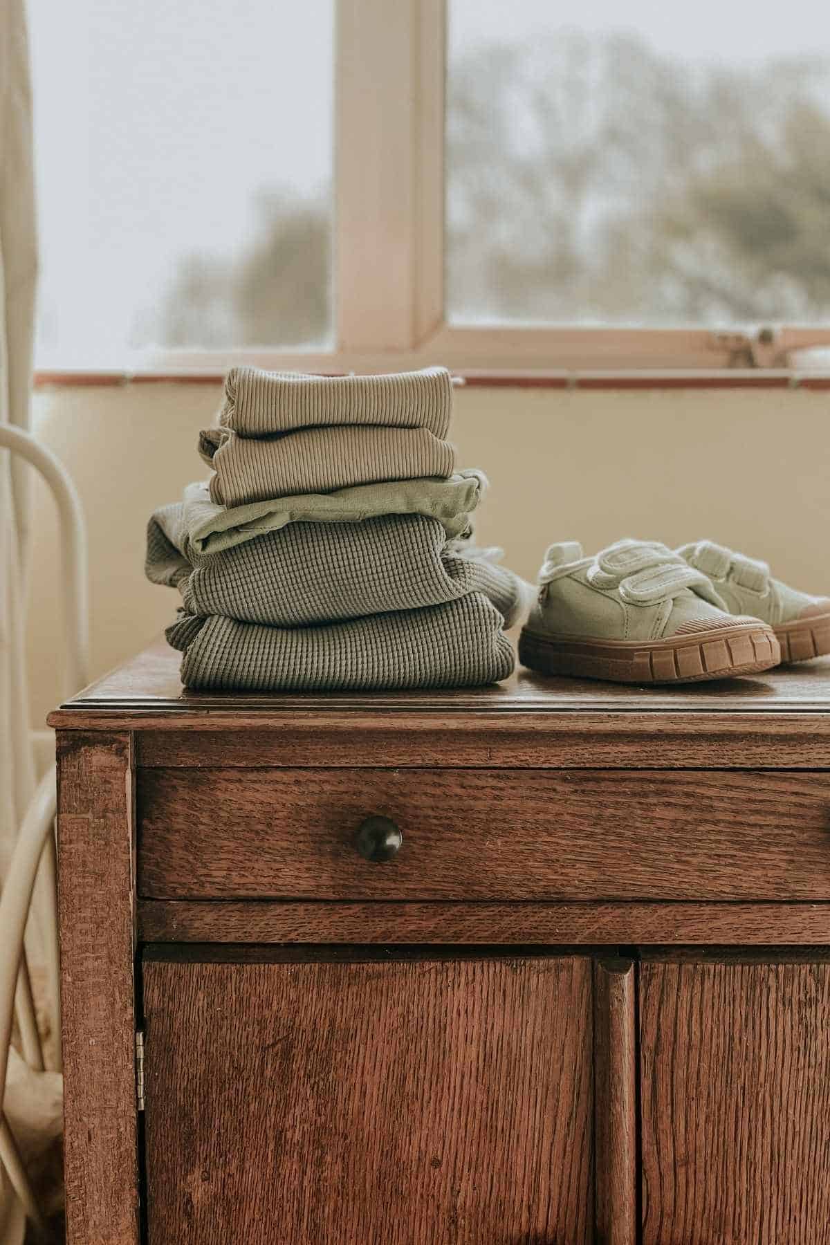 A wooden dresser with neatly folded green Kidly clothes and a pair of green shoes on top. A window in the background allows soft natural light to enter the room.