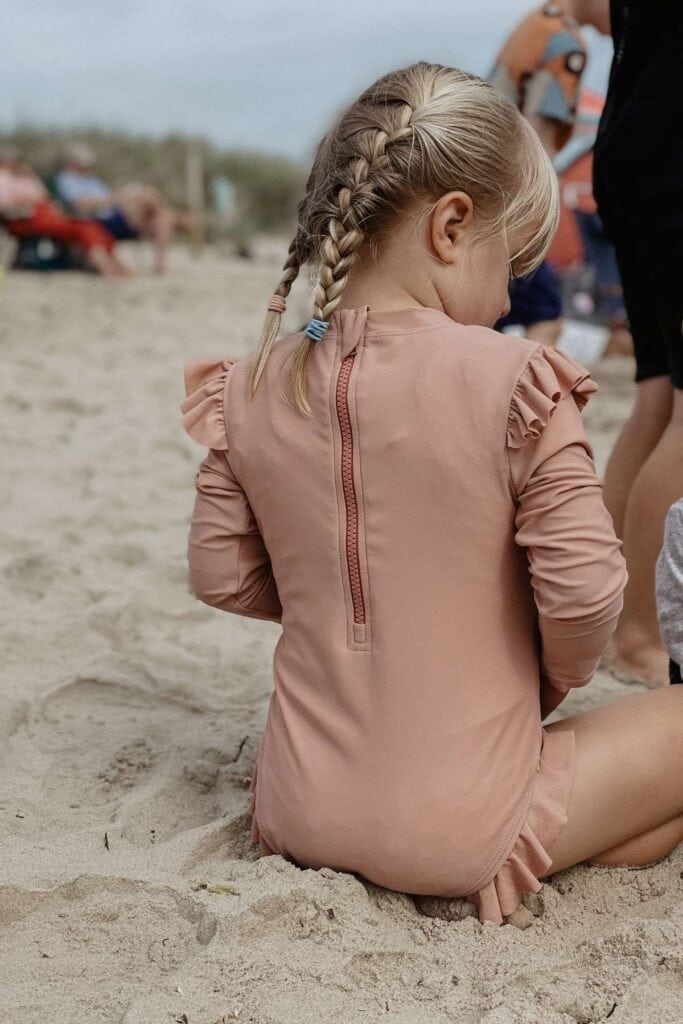 A young girl with braided hair sits on a sandy beach, wearing a pink Kidly swimsuit with ruffled sleeves. She is turned away from the camera, with people and beach gear visible in the background.