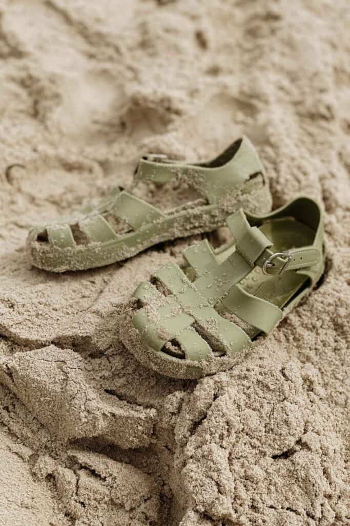 A pair of olive green Kidly jelly shoes with cut-out designs are placed on sandy ground, partially covered in sand. The sandals have a buckle strap and appear casual and relaxed in style.