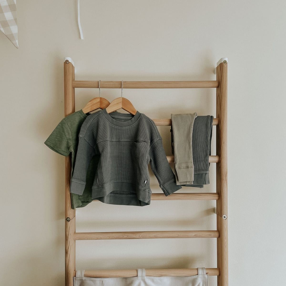 Kidly children's clothing displayed on a wooden ladder against a beige wall. The clothes include a green t-shirt and a long-sleeve gray top with matching pants. A folded blanket is at the bottom of the ladder.