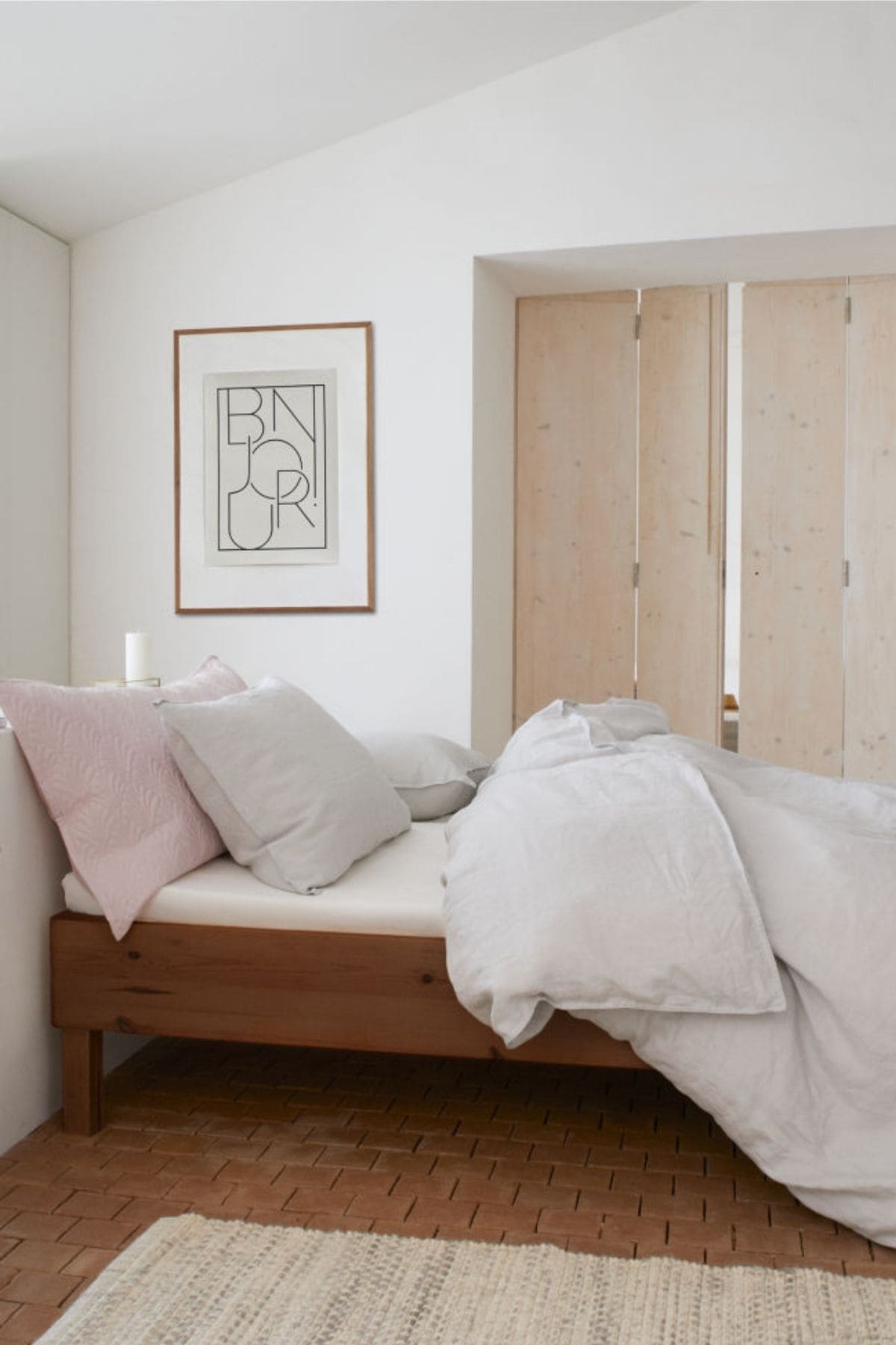 Minimalist bedroom with a wooden bed adorned with white and pink pillows and a white duvet with affordable linen sheets. There's a framed abstract art piece on the wall and beige wooden sliding doors. A textured light carpet is on the floor.