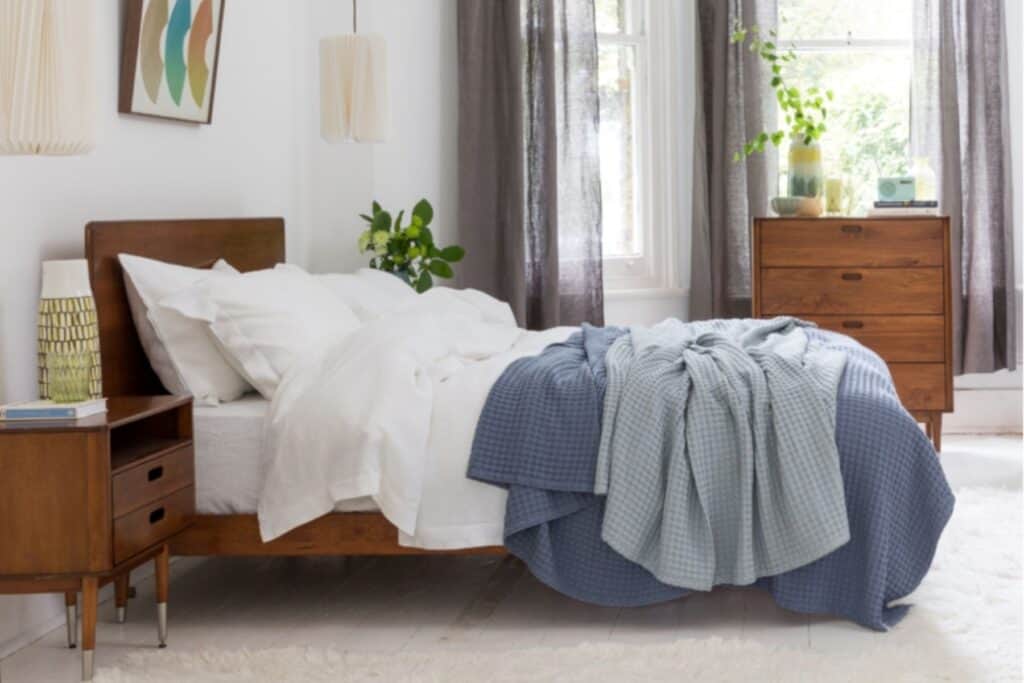 A cozy bedroom with a wooden bed frame and white linen bedding, partially covered by a blue textured blanket. A wooden nightstand and dresser flank the bed. Large windows with sheer curtains let in natural light, and potted plants add a touch of green.