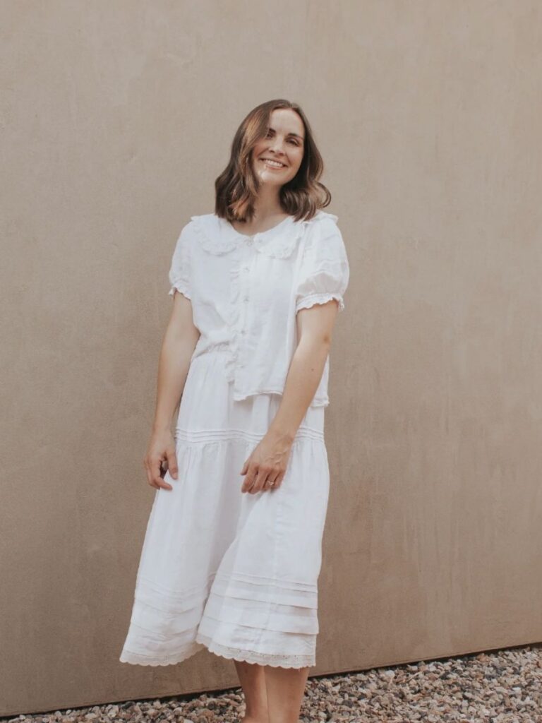 A woman in a white two-piece outfit with delicate lace details, featuring a ruffled blouse and a tiered, flowing Madswick Zamora skirt, evoking vintage and cottagecore aesthetics.