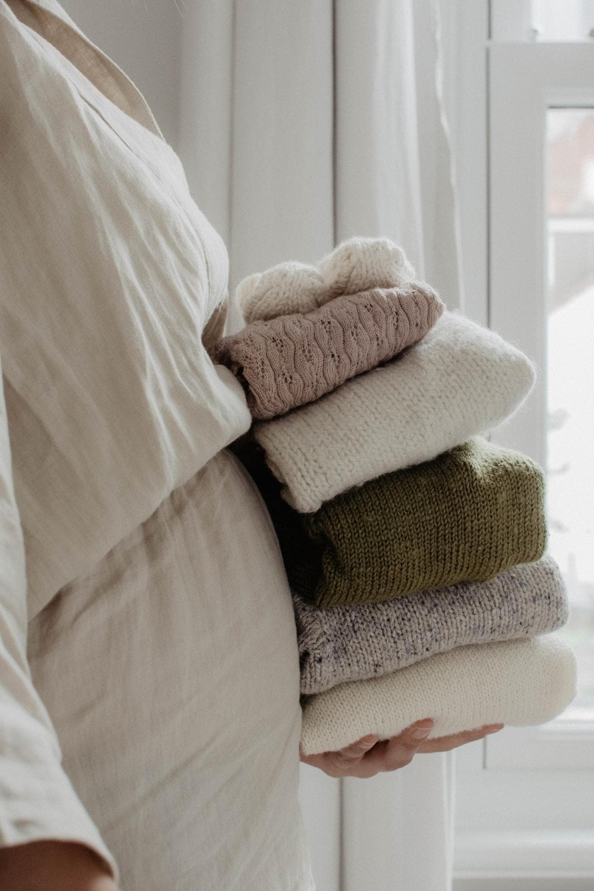 A person, comfortably draped in a light, flowing garment, holds a stack of neatly folded sweaters in neutral tones like beige, pink, green, and gray. They stand near a window with light curtains that let the sunshine gently glow around them.