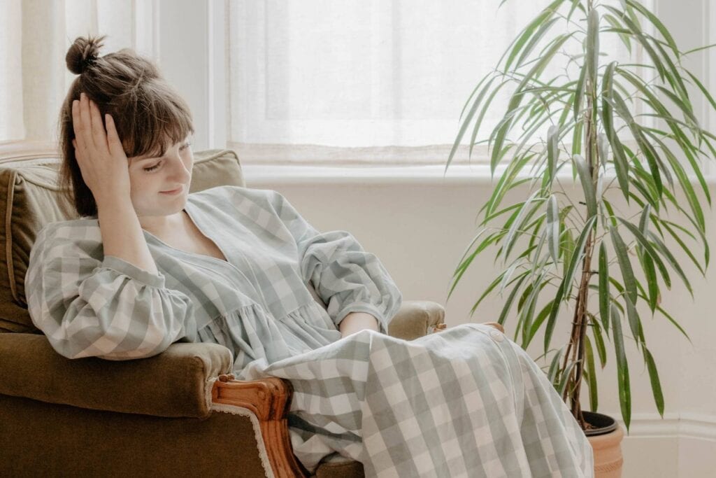 A person with a topknot hairstyle sits on a cushioned armchair, wearing a checkered dress that epitomizes stylish dressing. They rest their head on one hand, appearing relaxed. A tall potted plant is beside the chair, and sheer curtains allow soft light into the room.