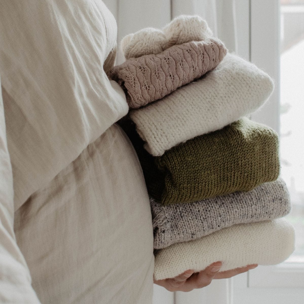 Person standing by a window with light filtering through sheer curtains, comfortably holding a stack of folded sweaters in neutral and green tones.