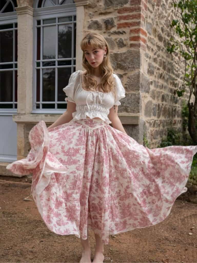 A woman in a billowing, vintage-inspired skirt and a ruched blouse, standing against a rustic stone building. The skirt, the Maria Juterud Darcy Skirt, features a gathered waist and a delicate toile print in soft pink and cream.