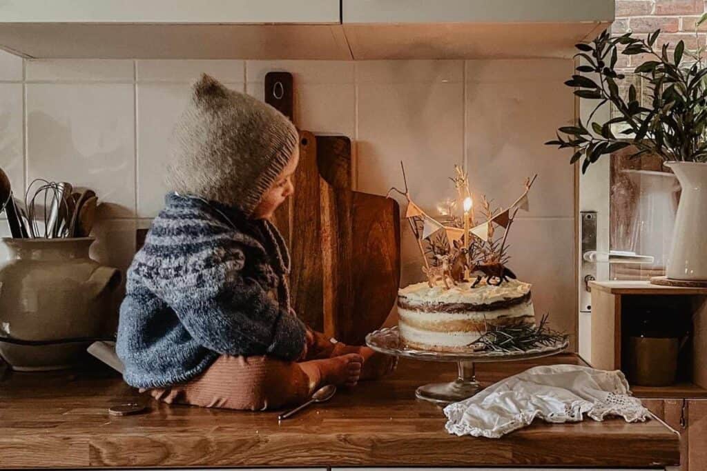 A small child wearing a cozy sweater and knit hat sits on a kitchen counter next to a decorated cake with lit candles. The scene is warmly lit, with kitchen utensils and a plant in the background, creating a rustic and cozy atmosphere.