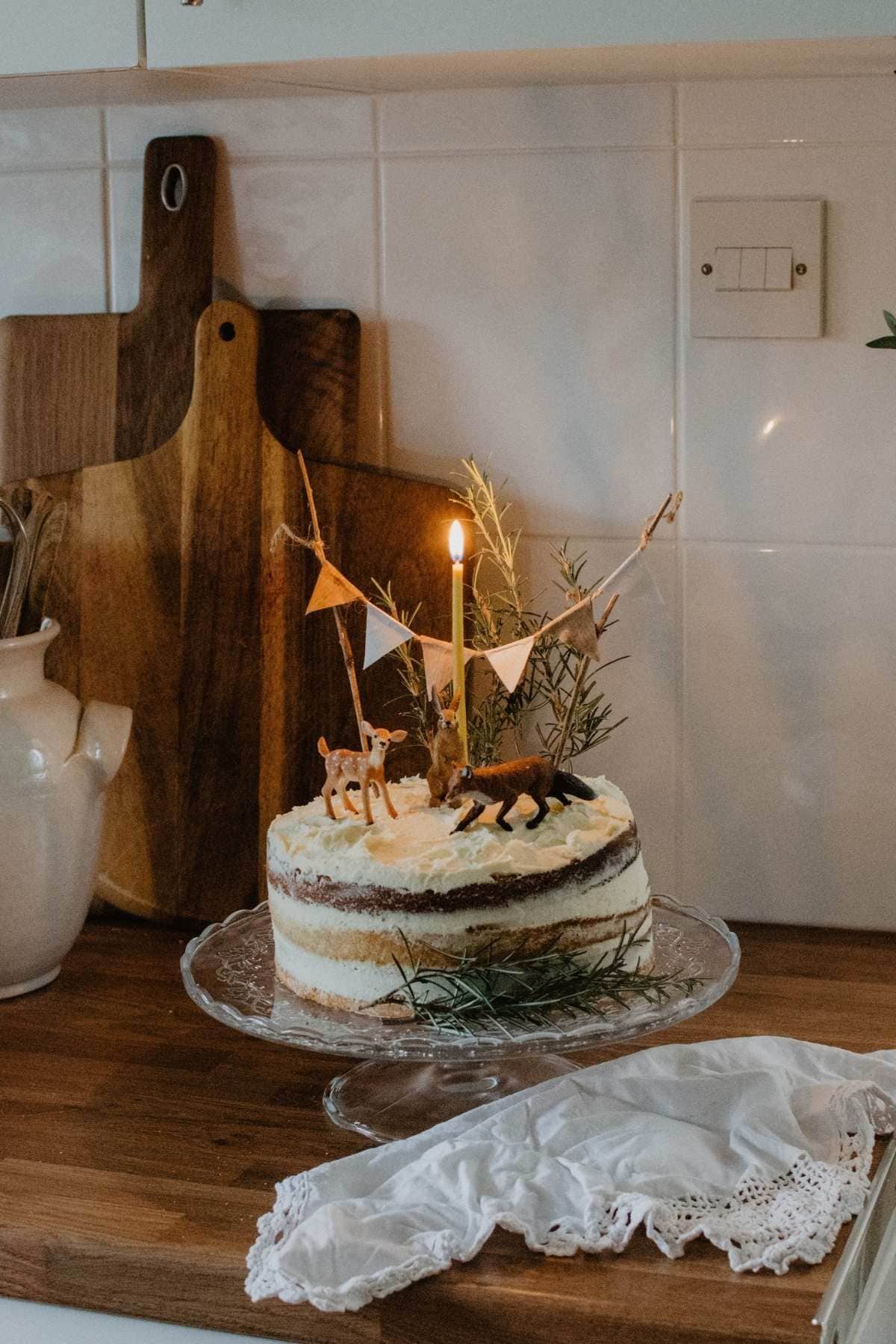 A rustic-themed naked cake on a glass stand is topped with small animal figures, a burning candle, and a banner. The cake sits on a wooden counter, surrounded by a white cloth and cutting boards against a tiled backsplash.