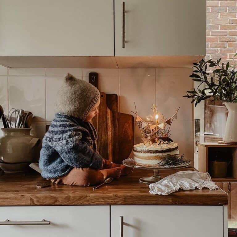 A small child wearing a knitted hat and sweater sits on a kitchen counter, gazing at a birthday cake with two lit candles. The kitchen is rustic with wooden accents and potted plants. A cozy, serene atmosphere surrounds the scene.