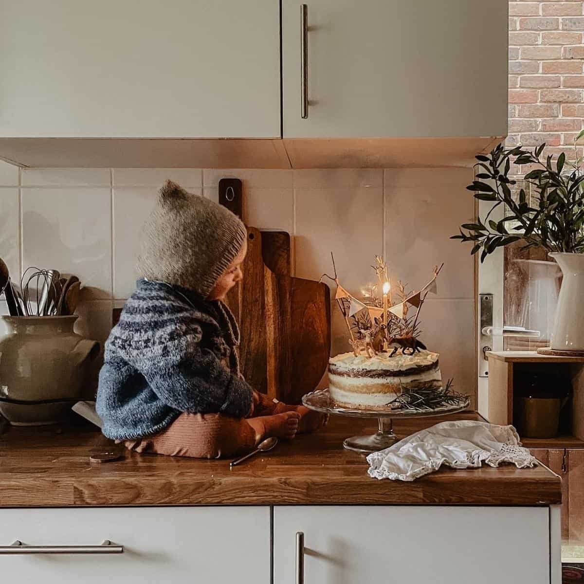 A small child wearing a knitted hat and sweater sits on a kitchen counter, gazing at a birthday cake with two lit candles. The kitchen is rustic with wooden accents and potted plants. A cozy, serene atmosphere surrounds the scene.