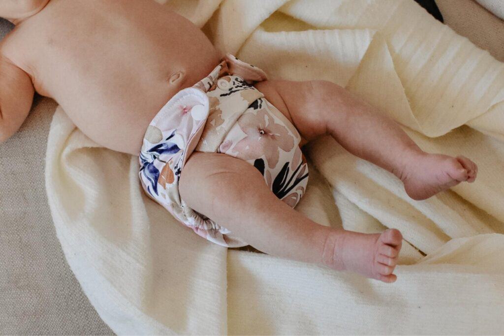 A baby lies on a soft beige blanket, showcasing a floral-patterned cloth diaper. With legs slightly bent, this scene highlights the comfort and style of newborn reusable nappies—a gentle introduction to choosing eco-friendly options for your little one.