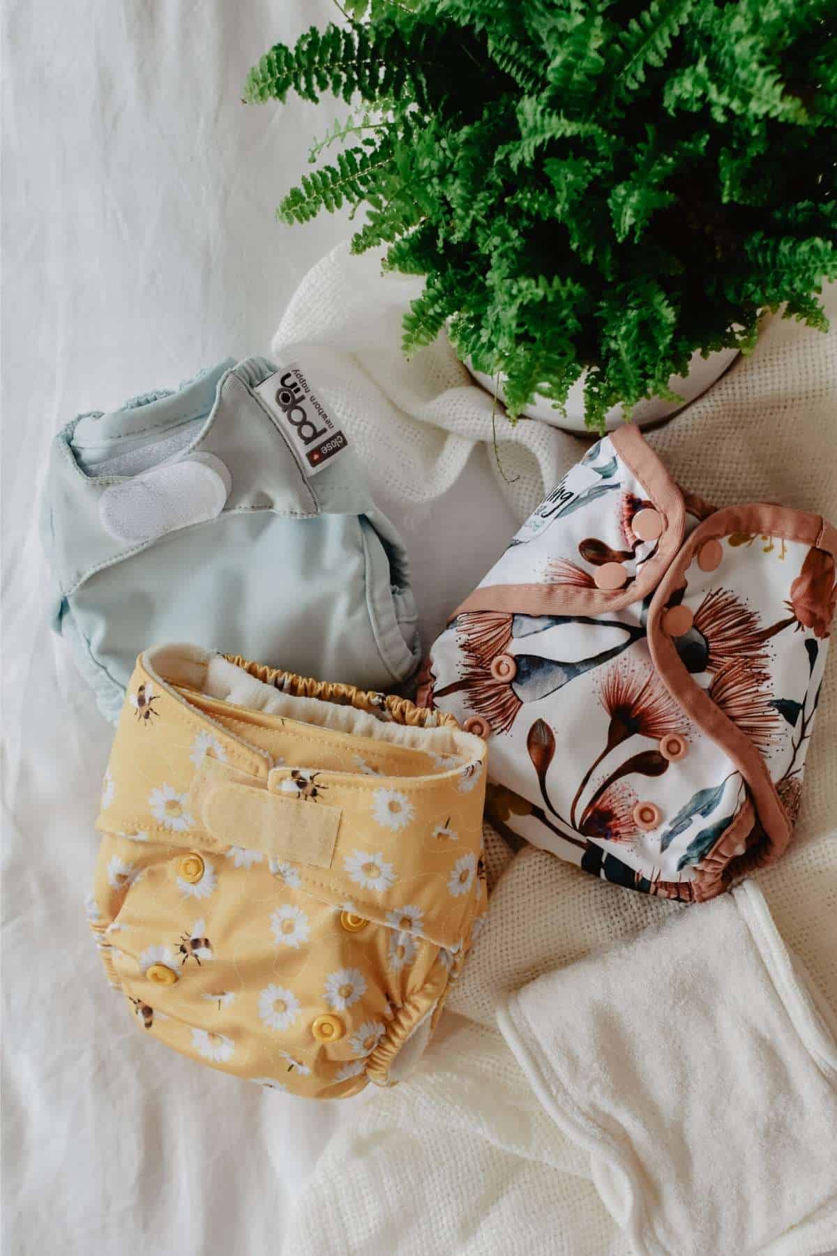 Three cloth diapers, perfect for newborn reusable nappies, are artfully arranged on a white cloth. One is light blue, another yellow with white flowers, and the third boasts a floral pattern of pink and brown accents. A green potted fern provides a serene backdrop.