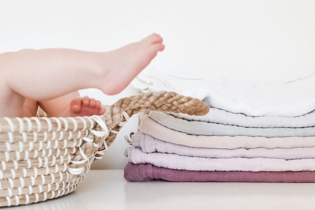 A babys feet playfully stick out of a woven basket on a white surface, while next to it, a neatly stacked pile of soft, pastel-colored muslin cloths in shades of white, pink, and lavender, which would make great nighttime reusable nappies