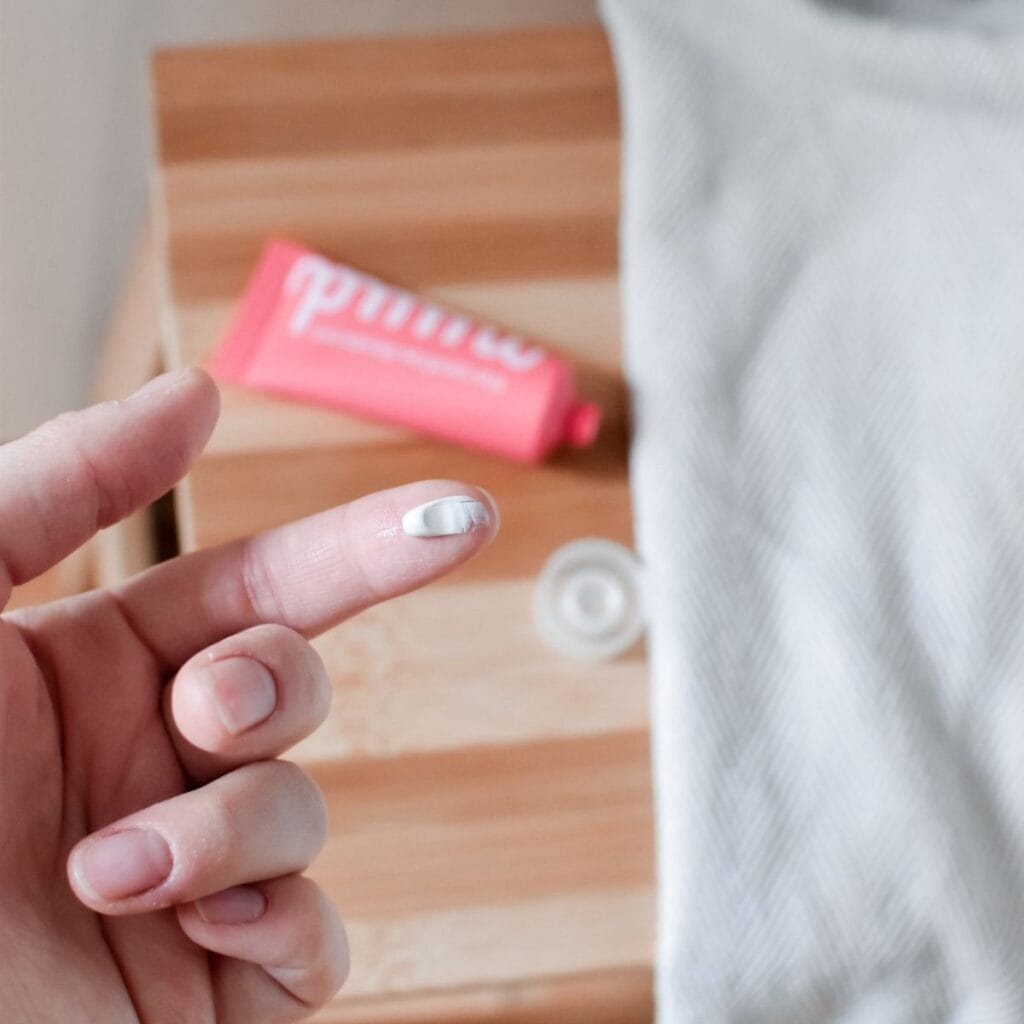 A hand with a finger extended shows a small amount of cream showing how much nuud deodorant you need to use each time. In the background, a wooden surface holds a pink tube labeled Nuud Deodorant and a white cloth. The tube features crisp white text.